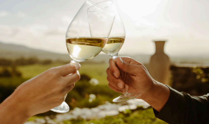 Duas mãos de pessoas brindam um vinho branco em uma taça de vidro.