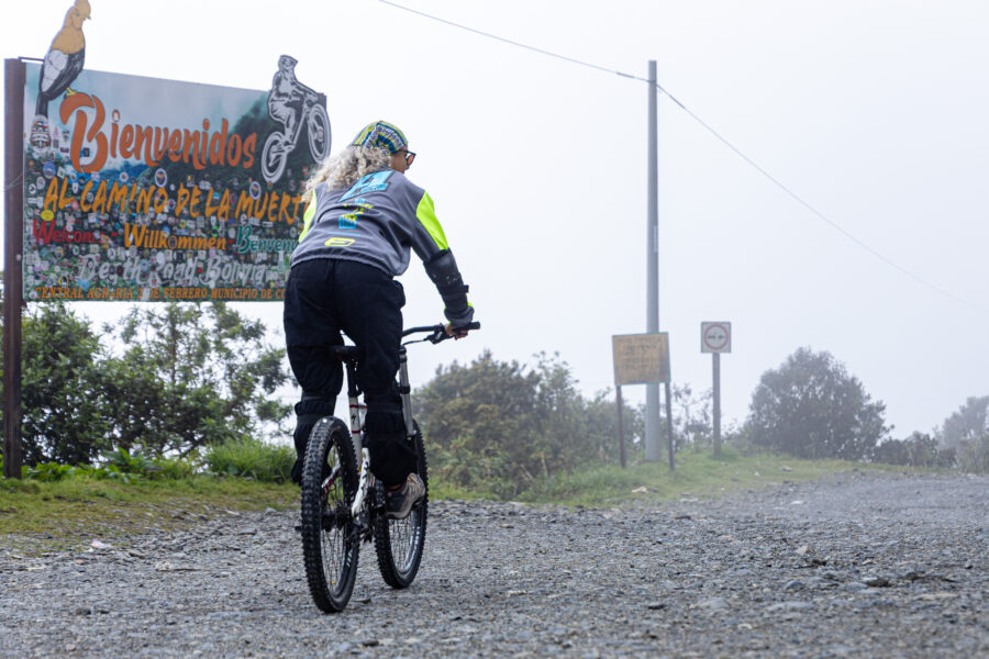 Agência de viagens ou viajar sozinho: Pessoa de costas andando de bicicleta em uma estrada de terra, passando em frente a uma placa de boas-vindas com a inscrição "Bienvenidos