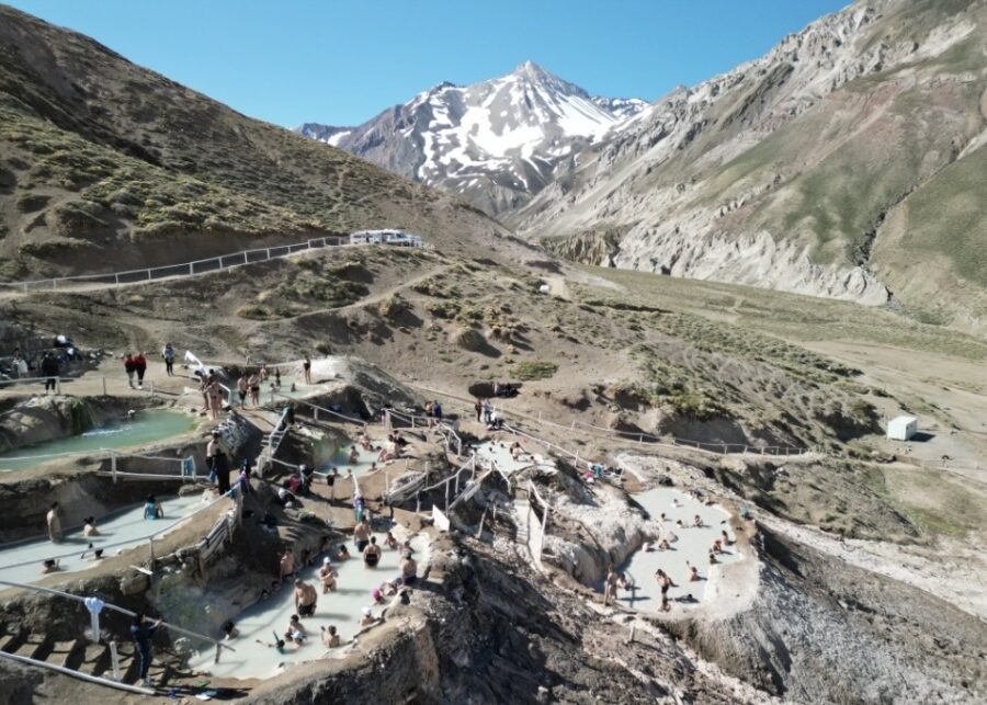 Vista ampla de piscinas termais naturais em um vale montanhoso próximo a Santiago, com várias pessoas relaxando na água.