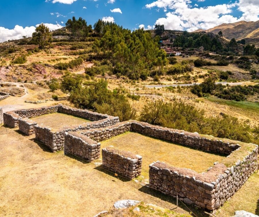 Ruínas de construções de pedra em área arqueológica cercada por vegetação e colinas, com estrada e pequenas casas ao fundo sob céu parcialmente nublado. Uma das aventuras que você pode viver com mais tranquilidade com seu glossário de viagem.