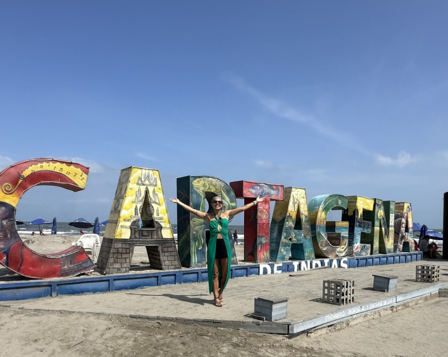 Pessoa posando em frente ao letreiro colorido “Cartagena”, próximo à praia, sob céu azul em um dia ensolarado na cidade colombiana.