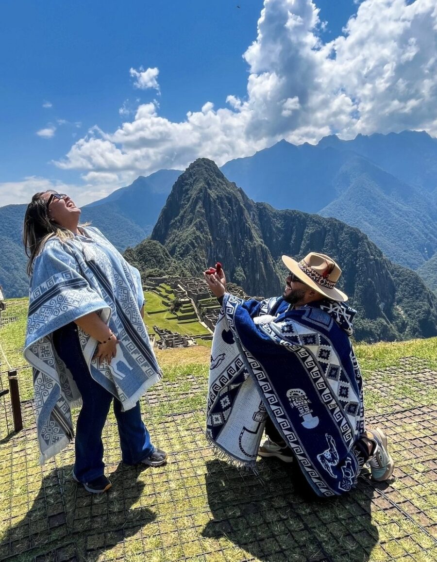 Homem ajoelhado pede alguém em casamento em um mirante com vista para ruínas de Machu Picchu e montanhas ao fundo, enquanto a outra pessoa reage emocionada.