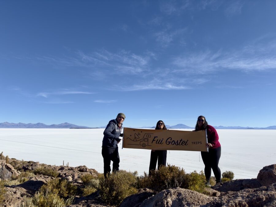 Três pessoas posam sorrindo em uma paisagem de deserto de sal sob céu azul, segurando uma placa escrita “Fui Gostei Trips”, com montanhas ao fundo. Cenãrio lindo para aproveitar com seu glossário de viagem.