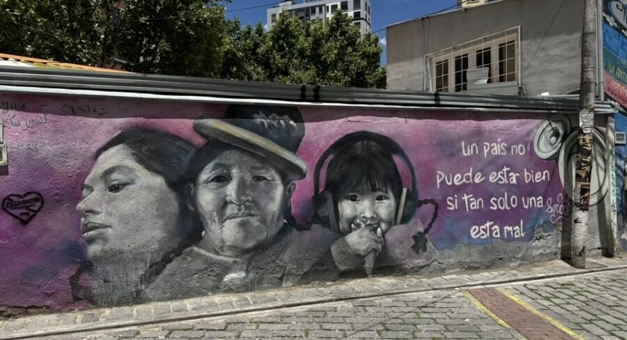 Mural de arte urbana com retratos de três pessoas — uma jovem, uma mulher idosa e uma criança com fones de ouvido — pintados em tons de preto e branco sobre fundo rosa, com frase em espanhol ao lado.