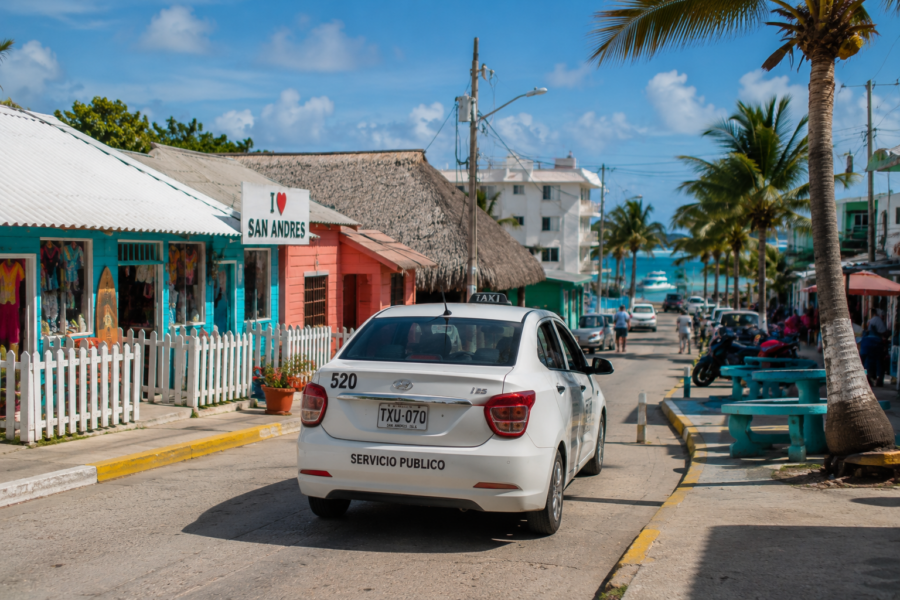 Táxi circulando por uma rua colorida de San Andrés, cercada por palmeiras, comércios locais e vista para o mar do Caribe ao fundo, representando uma das opções de transporte para quem busca como se locomover em San Andrés.