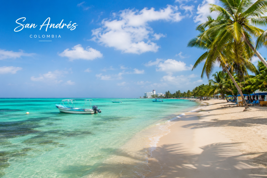 Vista de uma praia de San Andrés, na Colômbia, com águas em tons de azul-turquesa, areia clara, coqueiros à beira-mar e pequenas embarcações ancoradas próximo à costa.