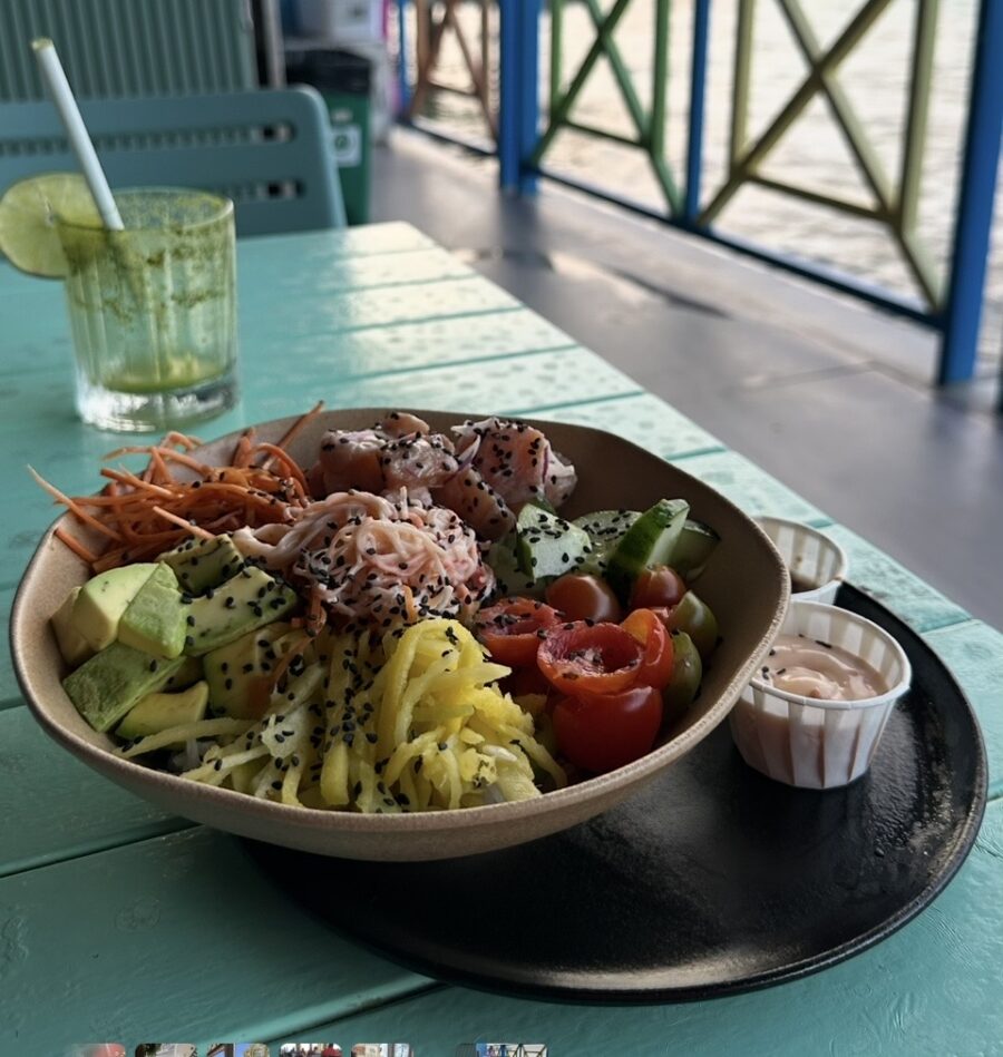 Poke bowl com cubos de peixe, abacate, pepino, tomate-cereja, manga, cenoura e molho, servido à beira-mar com uma bebida refrescante, destacando a variedade da gastronomia em San Andrés e as opções leves para um almoço com vista para o Caribe.
