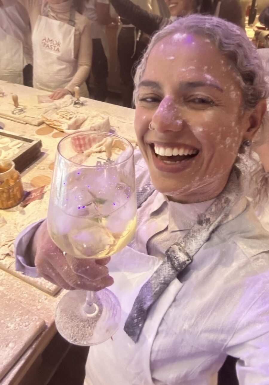 Pessoa sorrindo durante uma experiência no restaurante Amor e Pasta, segurando uma taça de bebida em um ambiente descontraído