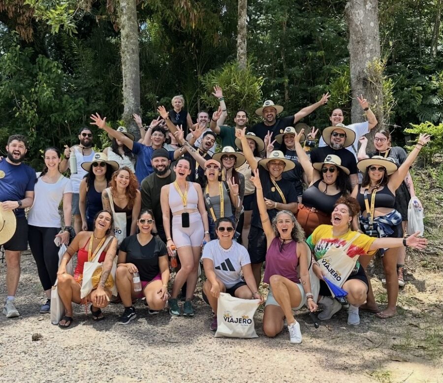 Uma foto em grupo vibrante ao ar livre com aproximadamente trinta participantes felizes da Fui Gostei Trips, posando na frente de árvores e vegetação exuberante. A maioria das pessoas está sorrindo ou fazendo o sinal de 'paz' e acenando, com muitas vestindo chapéus de sol e óculos escuros. Vários participantes seguram sacolas de lona estampadas com a marca "Viajero". A luz do sol incide sobre a cena