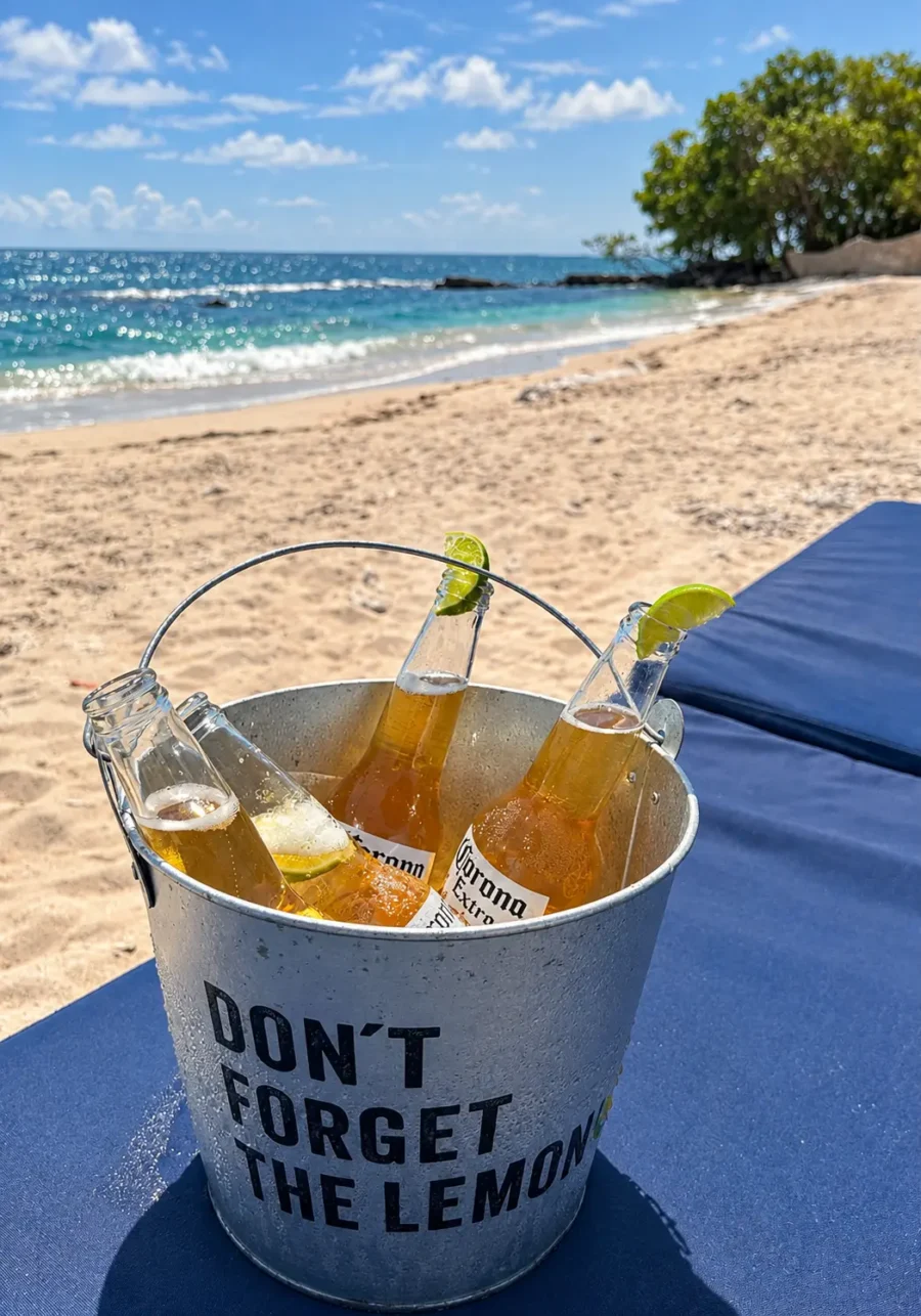 Um balde de gelo de metal contendo quatro garrafas de cerveja Corona Extra com rodelas de limão, apoiado em uma espreguiçadeira azul na areia de uma praia em Isla Corona em Cartagena. As garrafas estão geladas e com condensação. O texto "DON'T FORGET THE LEMON" está impresso em preto no balde. Ao fundo, vê-se o mar turquesa, a areia clara, algumas árvores e um céu azul com nuvens. O sol brilha intensamente