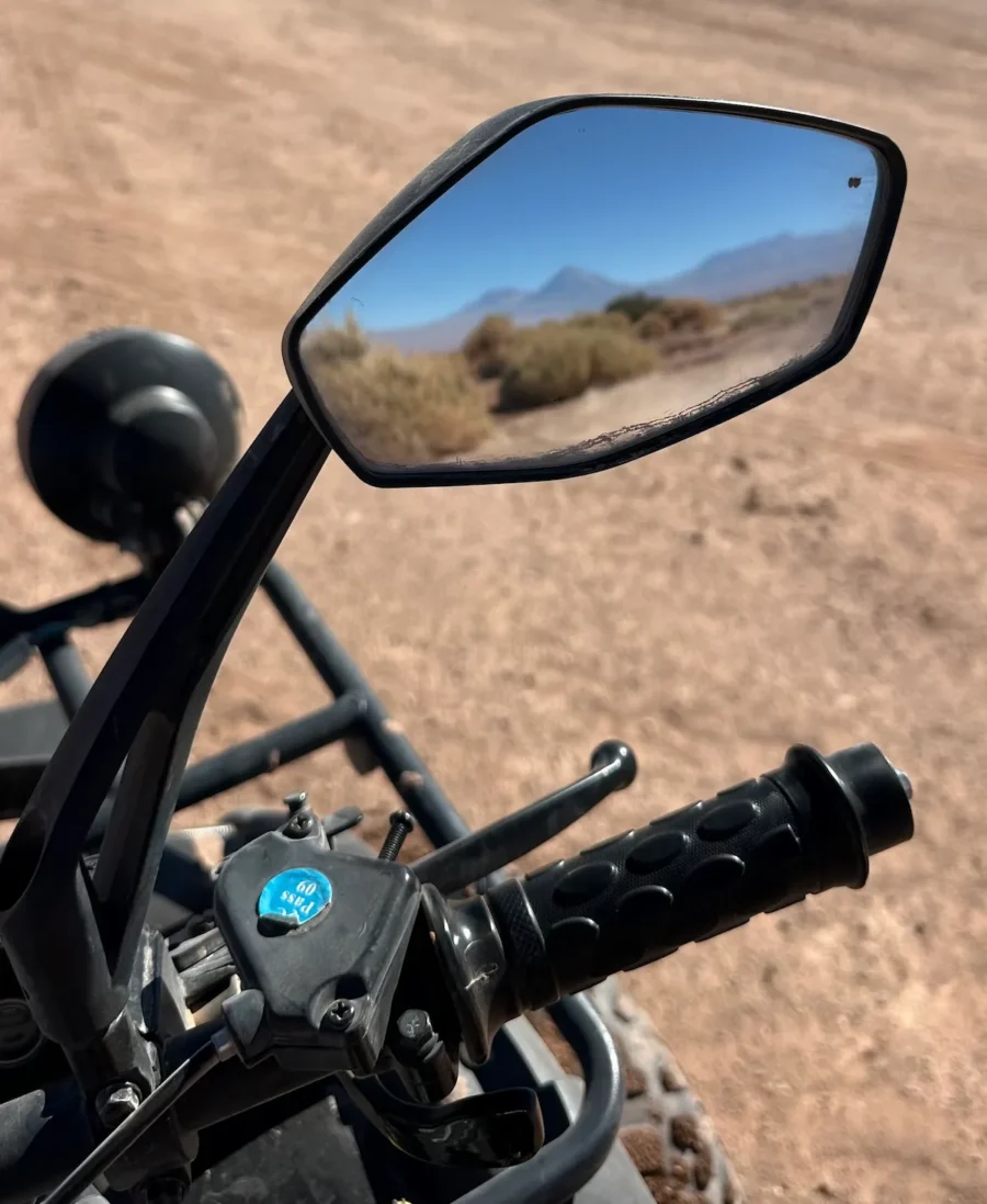 Detalhe do guidão e do espelho retrovisor de um quadriciclo durante um passeio de quadriciclo no Atacama, refletindo a paisagem desértica e as montanhas ao fundo