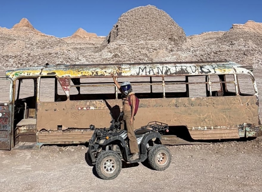 Pessoa com capacete de proteção em pé ao lado de um quadriciclo estacionado em frente à carcaça enferrujada de um ônibus antigo no deserto durante um passeio de quadriciclo no Atacama