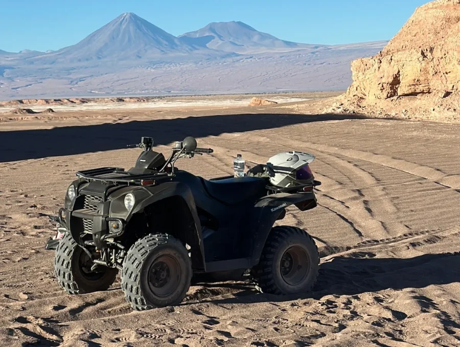 Quadriciclo preto estacionado na areia do deserto com um capacete apoiado no banco, tendo montanhas nevadas ao fundo em um passeio de quadriciclo no Atacama