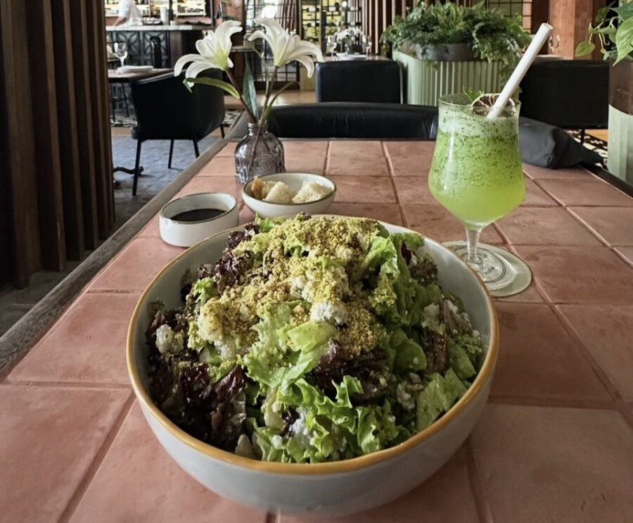 Uma tigela grande de salada verde com queijo esfarelado e castanhas, ao lado de um copo de suco verde, em uma mesa de azulejos terracota com lírios brancos ao fundo. A cena captura um momento da gastronomia em San Andrés