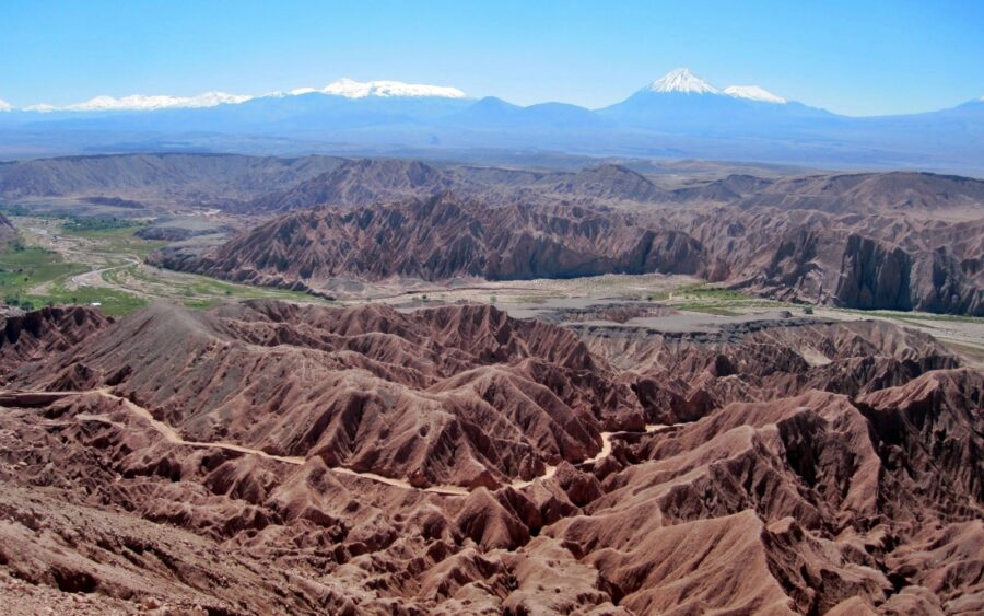 Como economizar no Atacama | Paisagem árida de alta altitude com rochas, solo seco e manchas de neve no primeiro plano, levando a vastos planaltos e vulcões distantes sob um céu azul claro