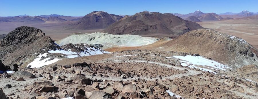 Como economizar no Atacama | Vista aérea e panorâmica das formações rochosas escarpadas e avermelhadas do Vale da Lua, com um vale verdejante ao longe e a cordilheira dos Andes nevada no horizonte