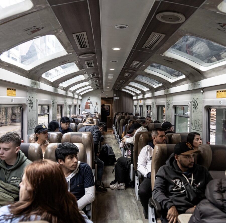 Interior de um vagão de trem moderno com teto solar panorâmico e grandes janelas. Passageiros sentados em poltronas de couro marrom estão olhando pela janela ou descansando. O corredor central é visível, com algumas pessoas em pé. O ambiente reflete a viagem confortável de um Machu Picchu bate-volta
