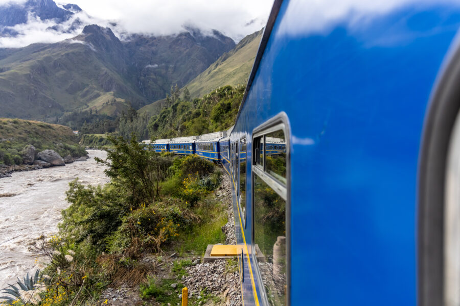 Uma vista lateral de um trem azul brilhante em movimento, com a câmera apontada para trás ao longo da lateral do trem. À esquerda, corre um rio caudaloso em um vale profundo e íngreme, cercado por montanhas verdes e rochosas, parte da rota cênica para um Machu Picchu bate-volta