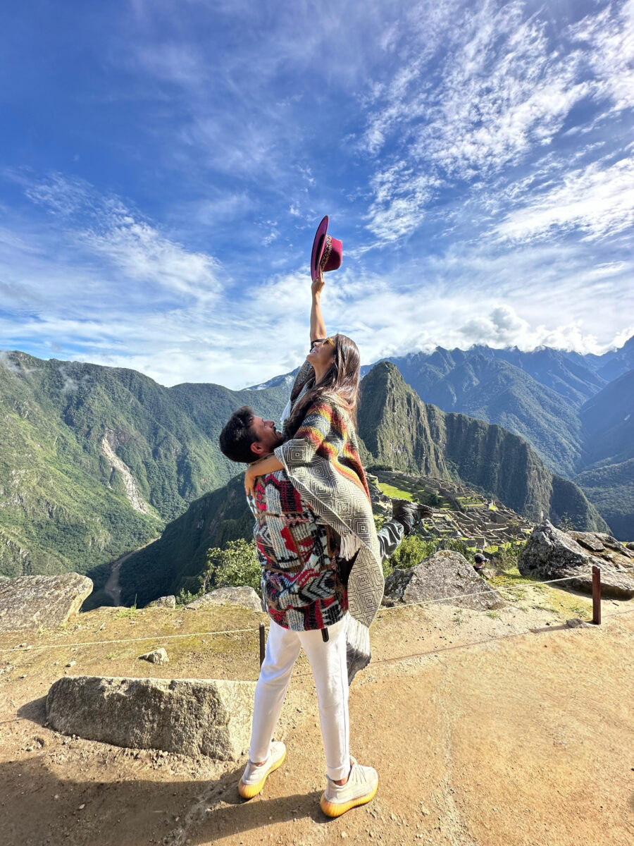 Um casal em pé nas ruínas de Machu Picchu, com o homem segurando a mulher no ar. A mulher, vestindo um poncho, levanta um chapéu roxo para o céu azul com nuvens brancas. Ao fundo, montanhas verdes e parte da cidadela inca, uma cena clássica de uma excursão de Machu Picchu bate-volta