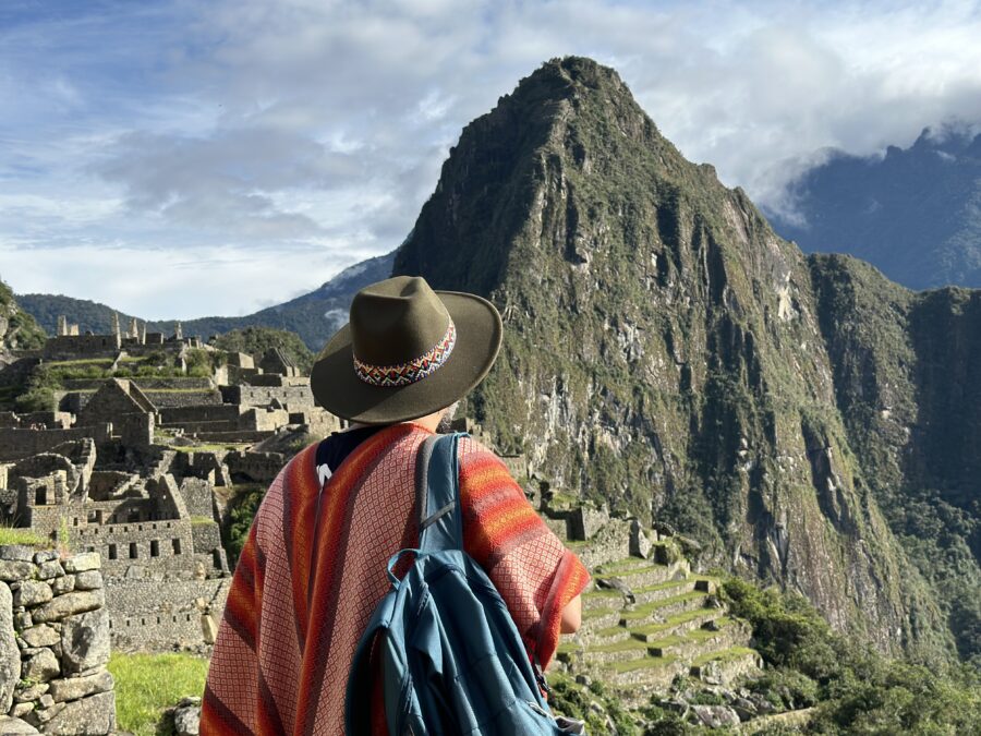 Uma vista por trás de uma pessoa usando um chapéu de feltro marrom com uma faixa colorida e um poncho xadrez laranja, segurando uma mochila azul, admirando as ruínas de Machu Picchu e o pico da montanha Huayna Picchu sob um céu parcialmente nublado. Este cenário é típico de uma viagem de um dia a Machu Picchu bate-volta