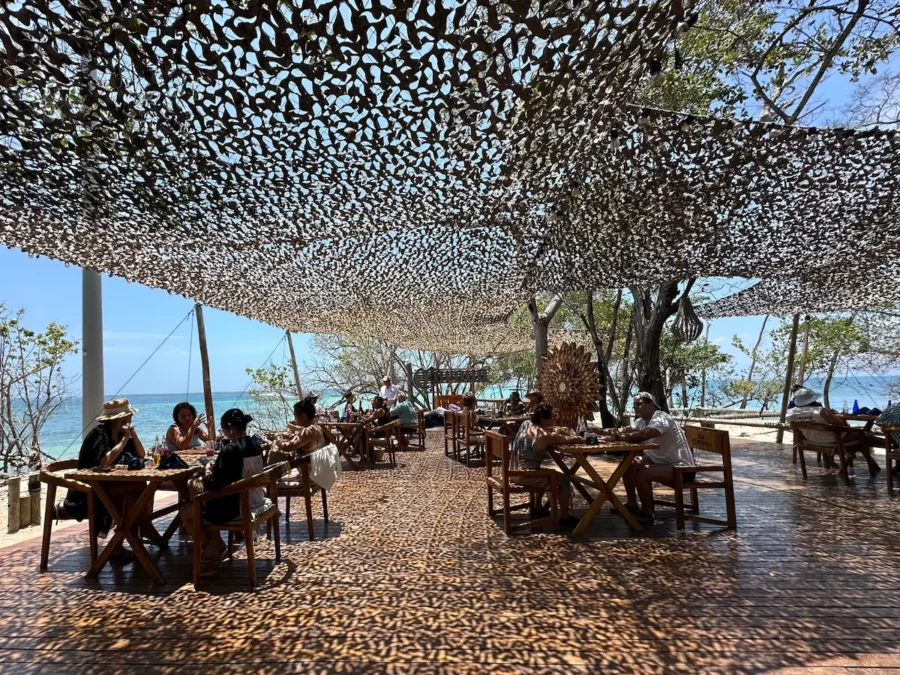 Uma área de jantar ao ar livre sombreada por uma rede de camuflagem na Isla Corona em Cartagena, Colômbia. Clientes sentam-se em mesas de madeira rústica em um deck de madeira texturizado. O mar azul-turquesa é visível ao fundo sob árvores maduras, criando uma atmosfera relaxante e sombreada
