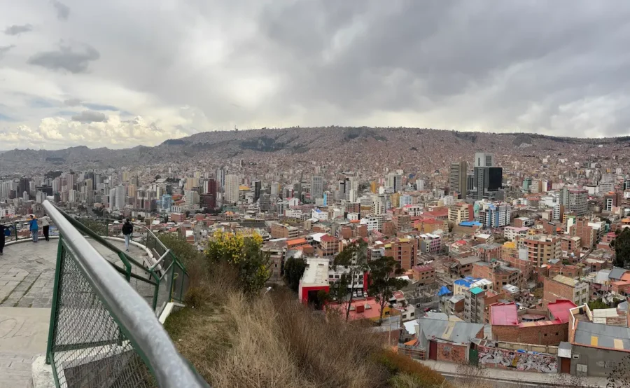 Vista panorâmica da cidade de La Paz, na Bolívia, a partir de um mirante elevado com uma grade metálica verde à esquerda. Vê-se uma vasta extensão urbana densamente construída que se estende por vales e colinas sob um céu nublado. Possibilidade de onde viajar no Ano Novo 2026 ou no Natal