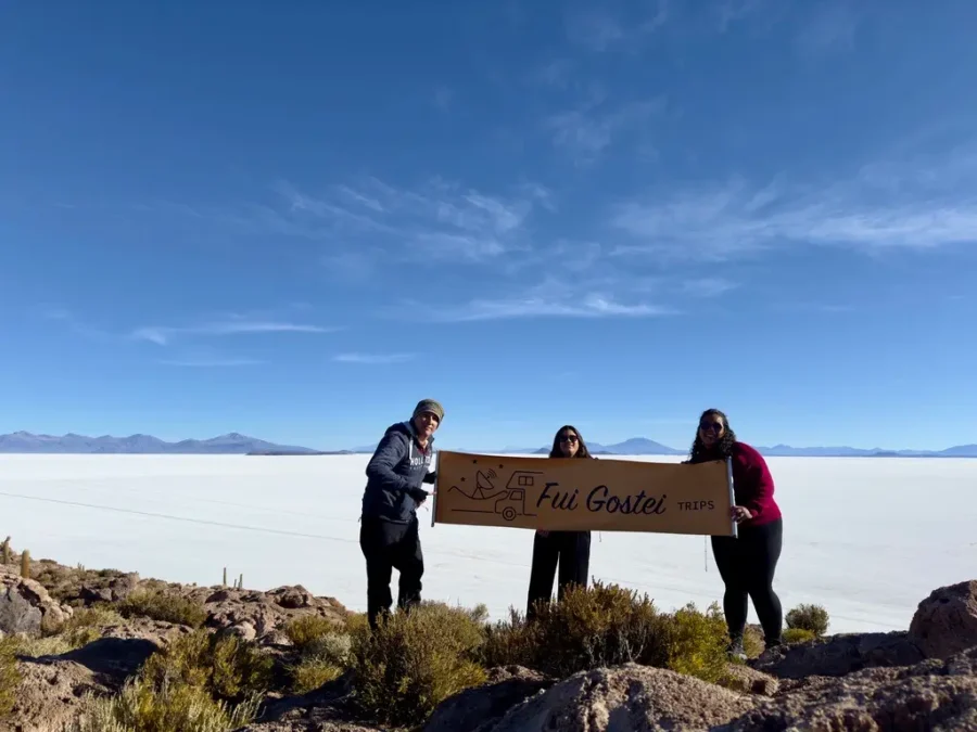 Três turistas em uma viagem em grupo, organizada por uma agência de viagens, posam segurando uma faixa no mirante do Salar de Uyuni