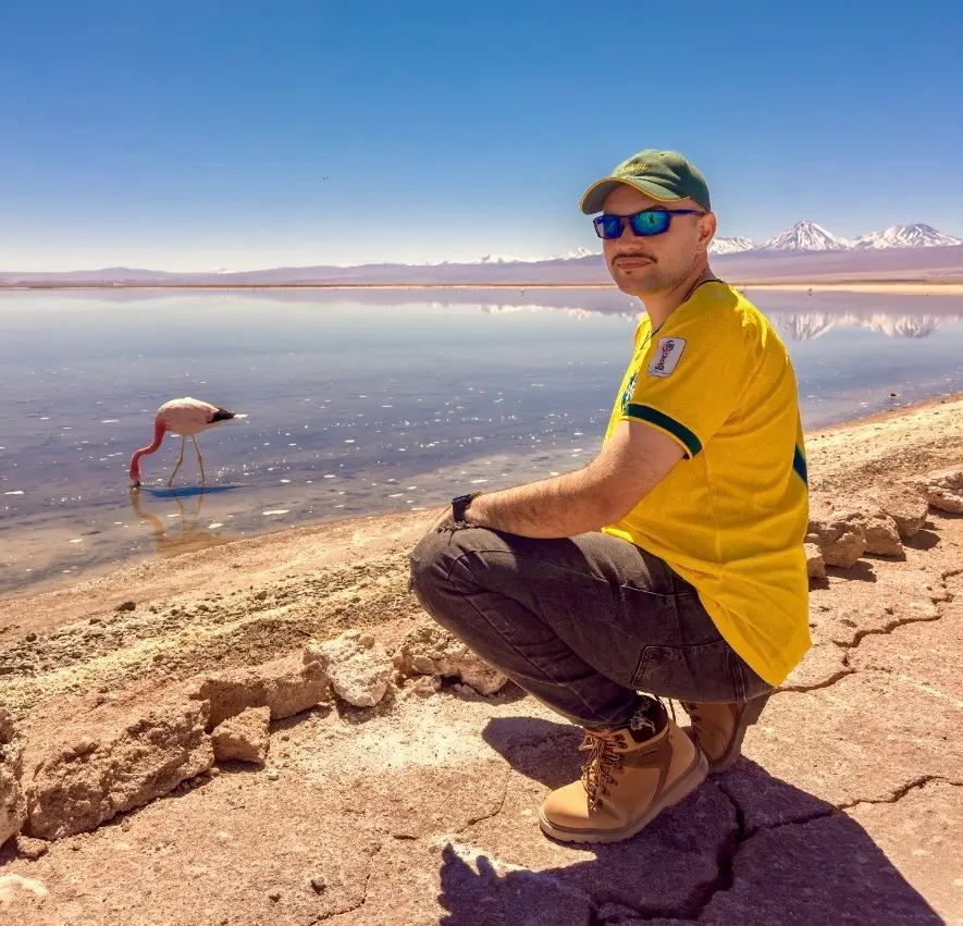 Salar de Uyuni Clássico, Conforto ou VIP: viajante usando camiseta amarela e óculos escuros agachado às margens de uma lagoa, ao lado de um flamingo, com montanhas nevadas refletidas na água